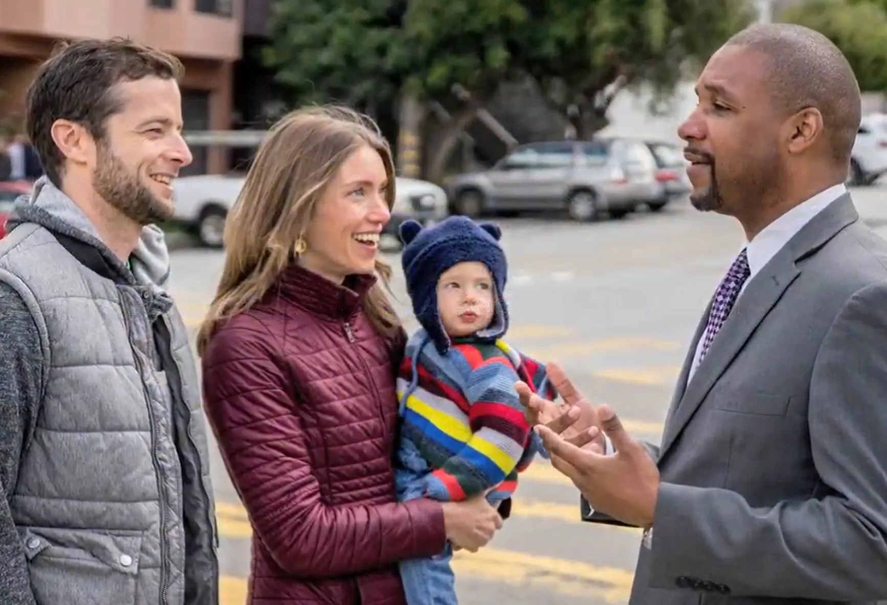 Candidate Shamann Walton at a street crossing, speaking with a smiling woman supporter holding a toddler in her arms