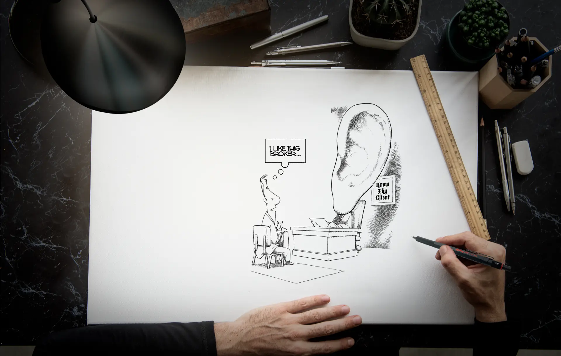 Artist's desk illuminated by a table-lamp, with artist's hands visible in frame as he draws an illustration using a Rotring pencil.